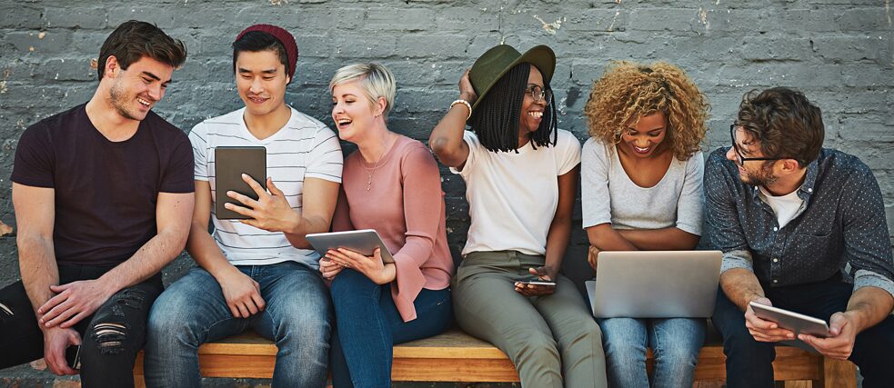Eine Gruppe junger Leute sitzen nebeneinander auf eine Bank. Sie lesen von Laptops und Tablets.