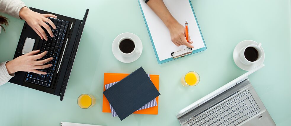 Draufsicht auf einen Tisch mit Laptops, Notizblocks, Schreibgeräten und Kaffeetassen