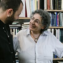 A young man with a black shirt, short hair and beard, and a middle-aged woman with short gray hair and a white shirt stand in front of bookshelves and among books. They look at each other and smile.