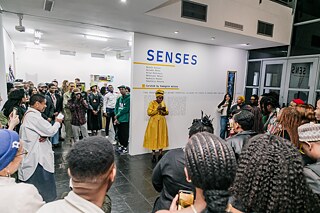 Das Bild zeigt eine Ausstellungseröffnung der Galerie. Eine Person in einem gelben Kleid steht im Mittelpunkt und spricht zu einer Gruppe von Menschen, die um sie herumstehen und zuhören. Im Hintergrund ist eine Wand mit dem Titel "SENSES" und einer Liste von Namen zu sehen. Die Galerie ist gut beleuchtet, und es sind viele Besucher anwesend, die aufmerksam der Rede folgen.