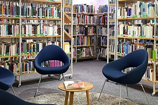Vor zwei Bücherregalen stehen in einem Kreis gestellt, vier blaue stoffbezogene Stühle um einen kleinen Tisch. Auf dem Boden liegt grauer Teppich.