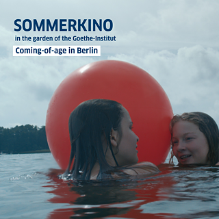 Two girls are in the water, playing with a large red inflatable ball on a sunny day.